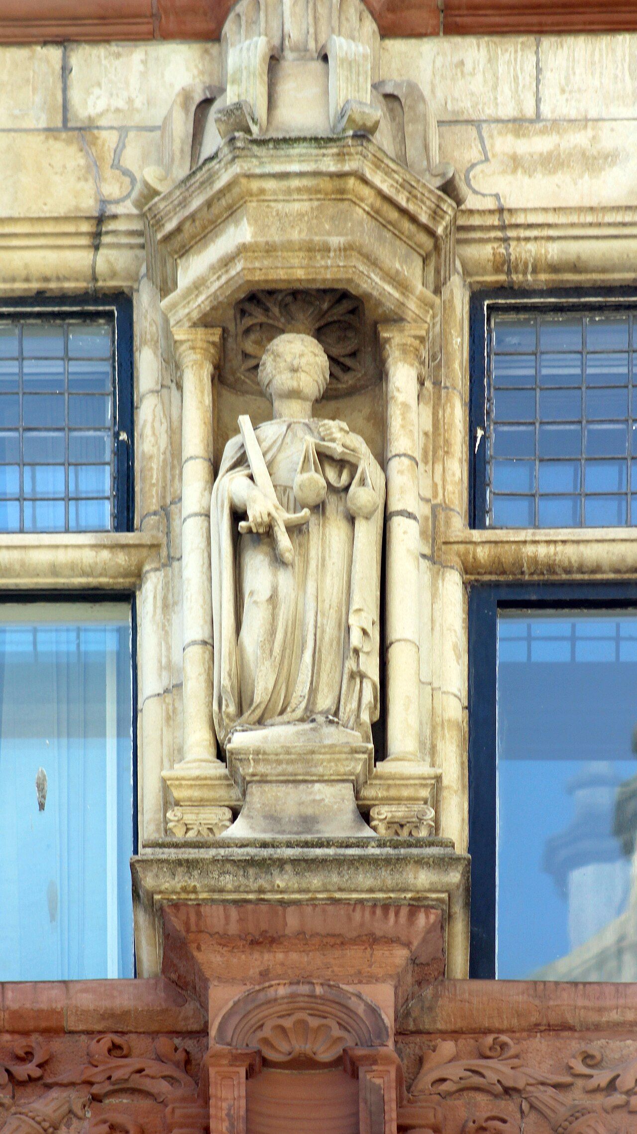 A statue of Justice holding balanced scales, blindfolded, against a stone courthouse fa&ccedil;ade.