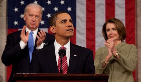 A speaker at a lectern addressing a large chamber, hands resting out of view on the lectern, with two seated officials applauding behind him.