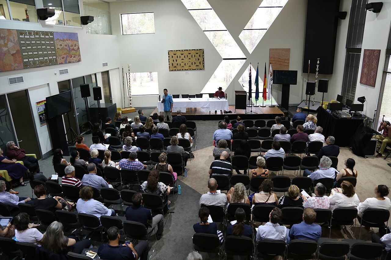 A speaker addresses a seated audience at a public meeting, with listeners showing varied expressions