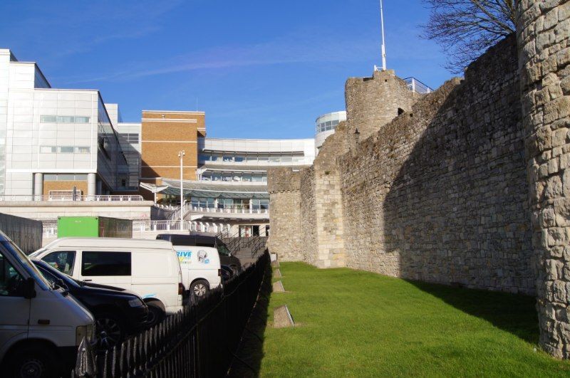 Colour photograph of a surviving stretch of Southampton's medieval stone town wall, with modern buildings, parked vans and railings in front of it